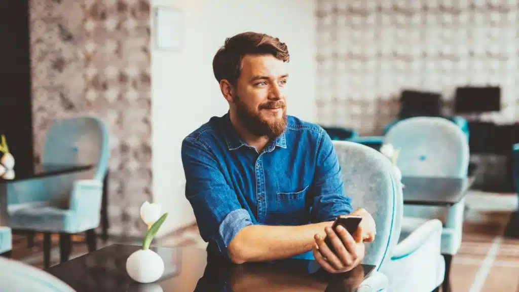 A man with a beard sits at a café table holding a smartphone and looking away.