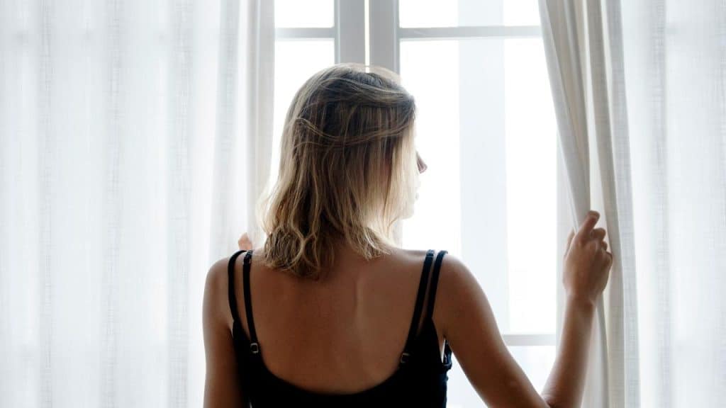 A woman standing by a window holding open sheer curtains.