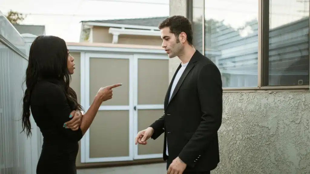 A woman in a black dress points her finger toward a man standing outside a building.