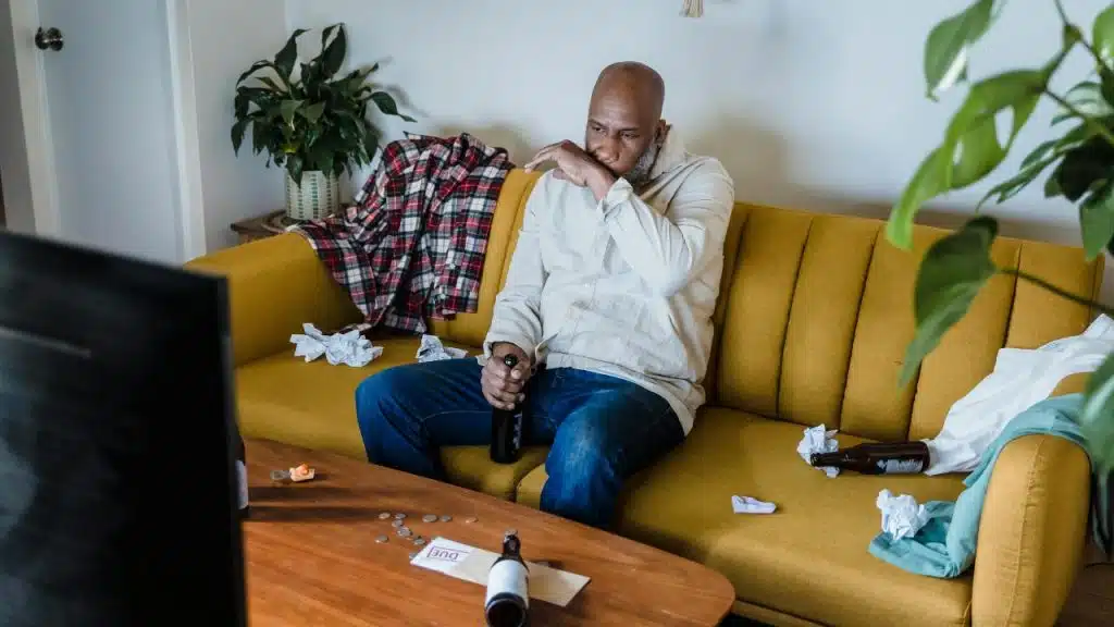 A man sits on a messy yellow sofa with bottles, papers, and clothes scattered around.