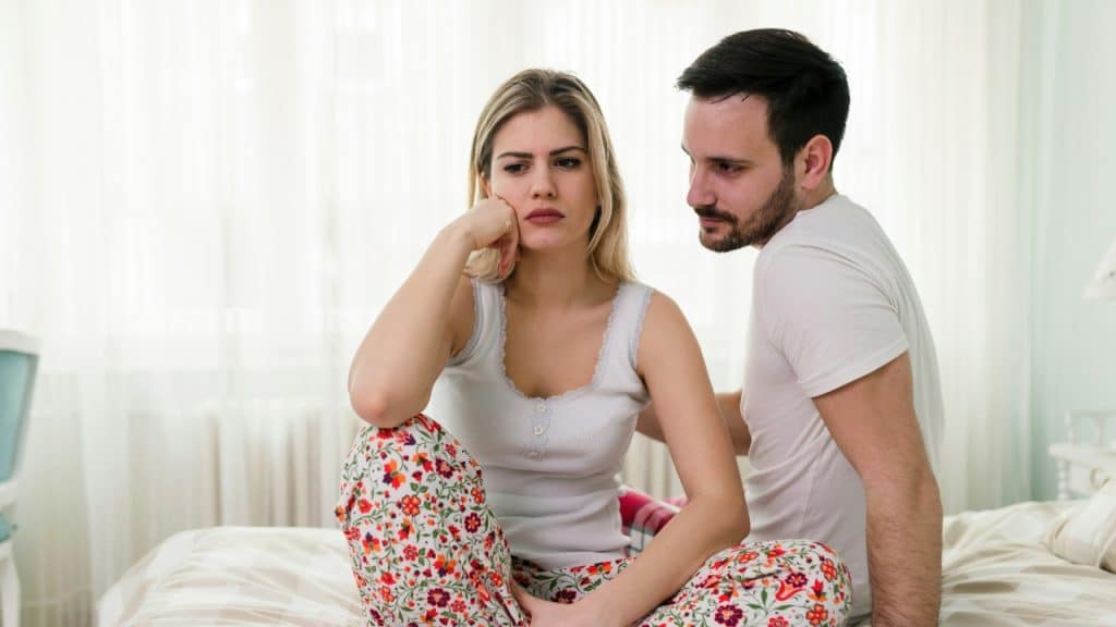 A woman in floral pants sits on a bed while a man looks at her.