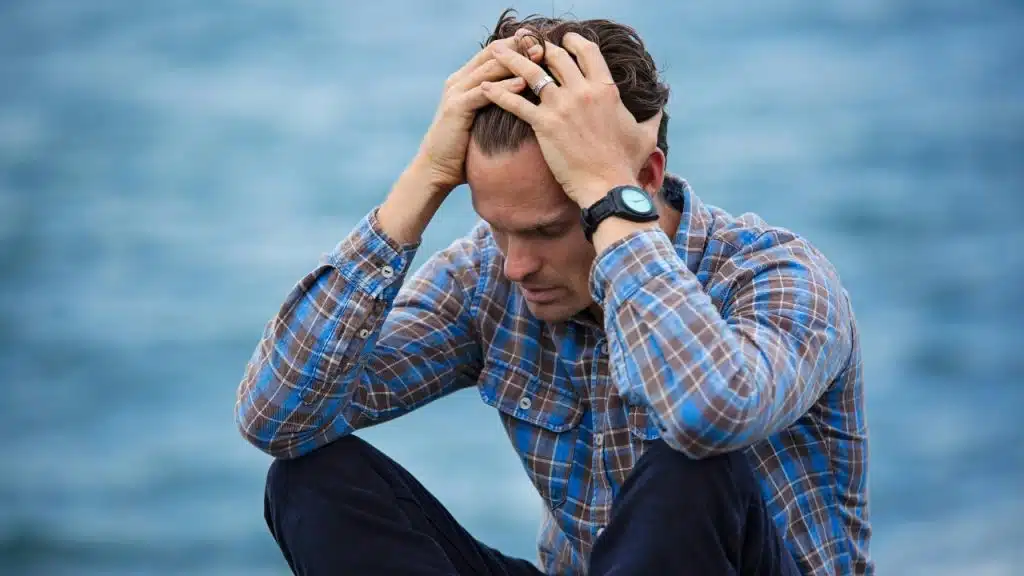 A man in a plaid shirt sitting by water with both hands on his head.