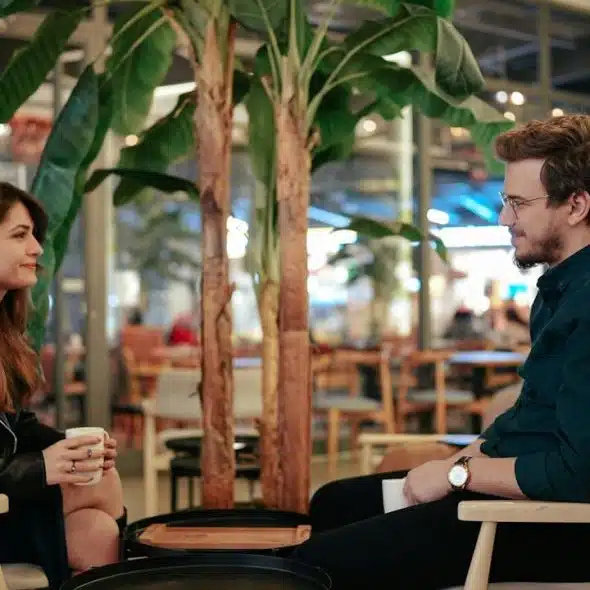 A couple is seated, facing each other in a brightly lit cafe or indoor seating area with plants, having a conversation over coffee.