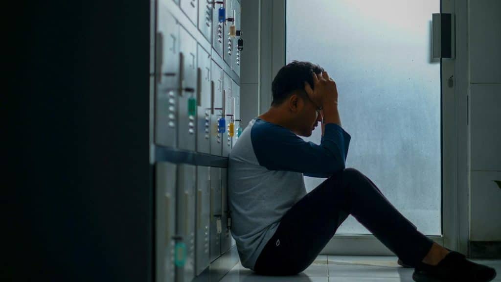 A man sitting on the floor while leaning on some lockers while holding his head in his hands