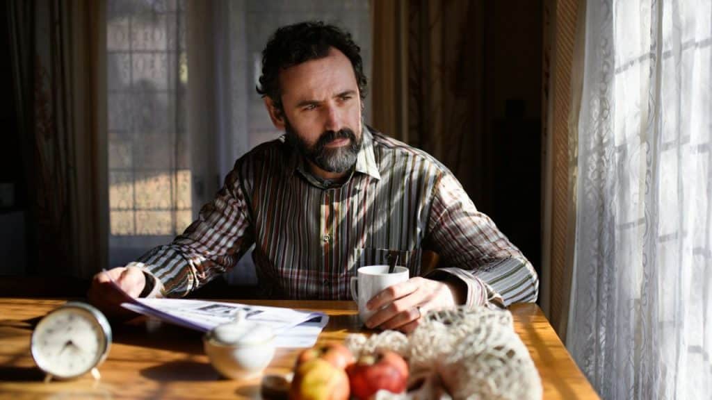 A bearded man in a striped shirt sits at a sunlit table with a newspaper.