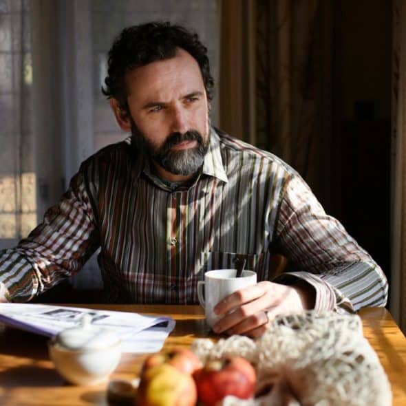 A bearded man in a striped shirt sits at a sunlit table with a newspaper.