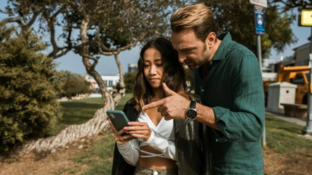 A man points at a phone held by a woman while standing together in a park.