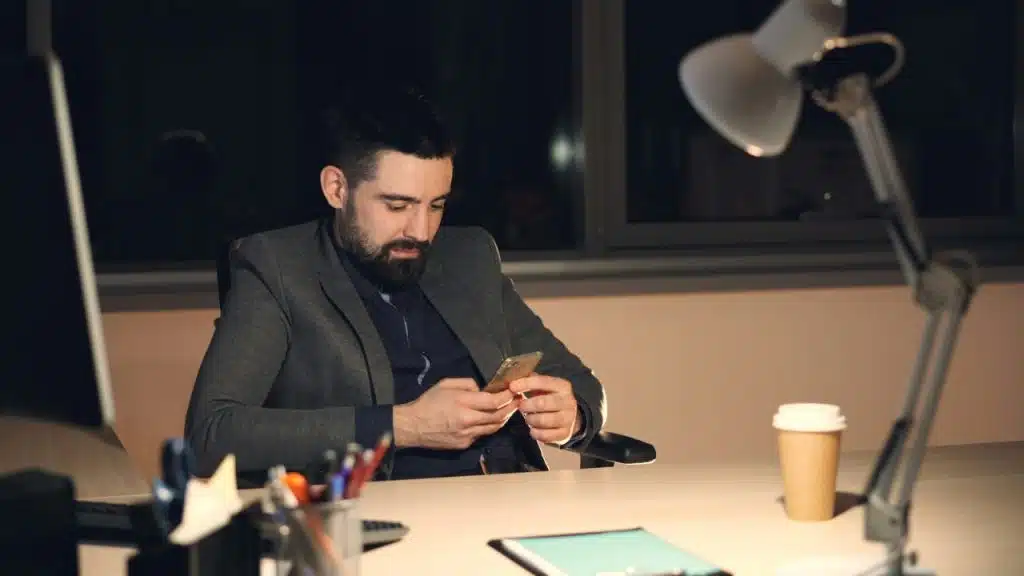 A man in a suit sits at a desk at night looking at a smartphone.