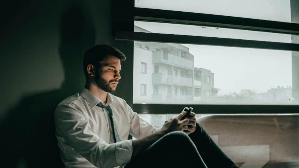 A man sitting on a window sill looking at his cell phone.