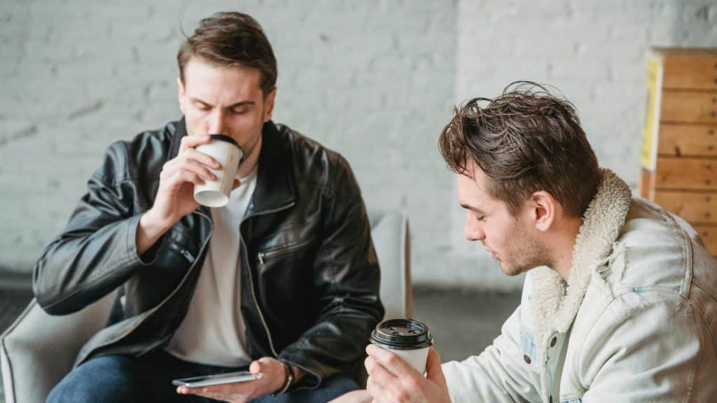 Two men sit in chairs holding coffee cups while one person looks at a phone.