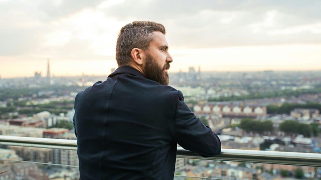 A bearded man in a dark jacket looks out over a city skyline from a balcony.