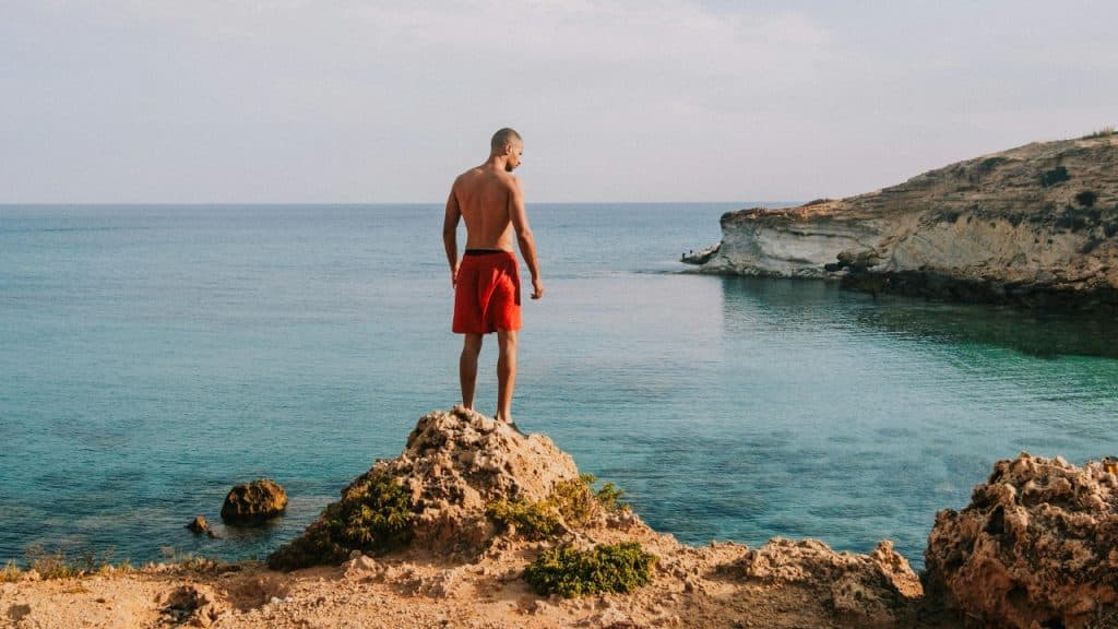 A shirtless man in red shorts stands on a rocky outcrop overlooking calm blue water.