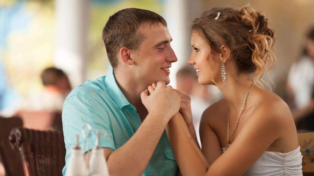 A man and woman in a restaurant hold hands and smile at each other.