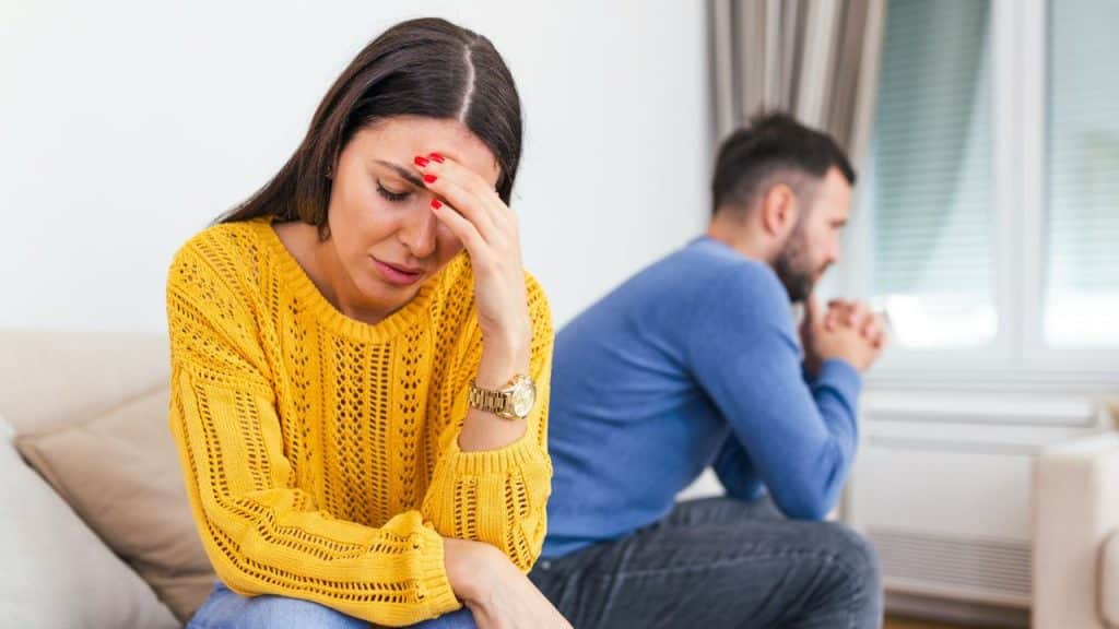 A woman in a yellow sweater touches her forehead while a man sits behind her.