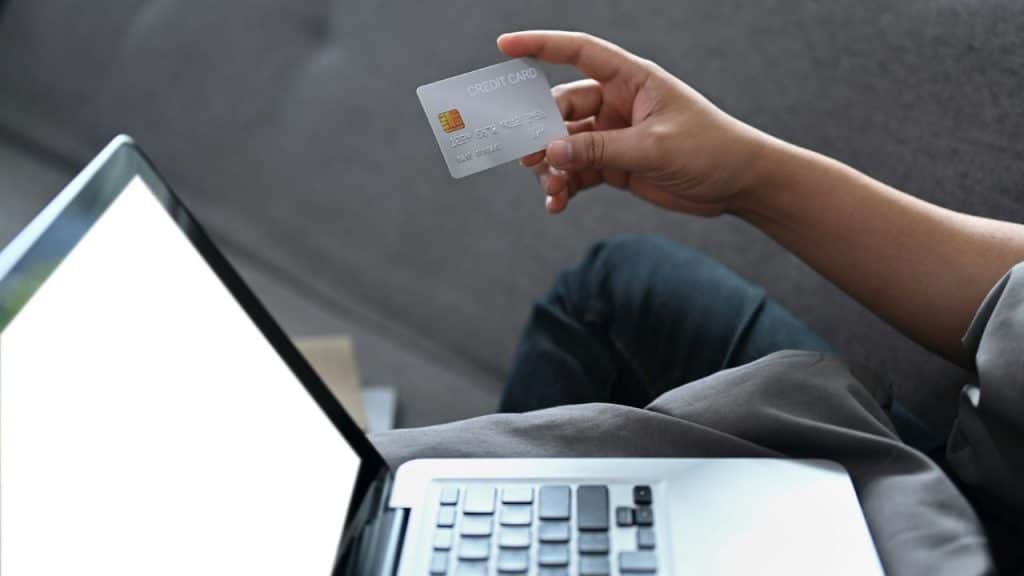 A person holds a silver credit card over an open laptop on a grey sofa.