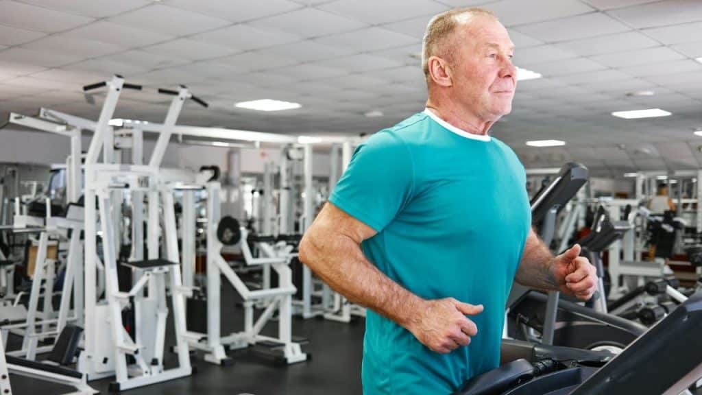 An older man in a teal shirt runs on a treadmill inside a gym with machines.