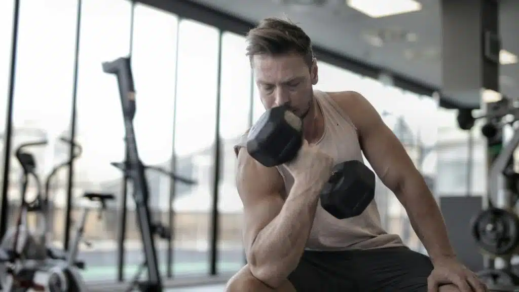A man in a tank top sits in a gym and lifts a black hex dumbbell.