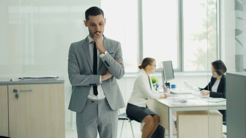 A man in a suit looks thoughtful while two women work at desks behind him.