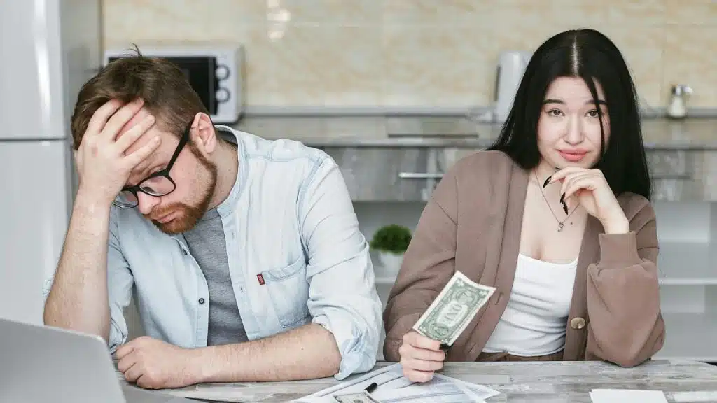 A man holds his head while a woman holds a dollar bill.