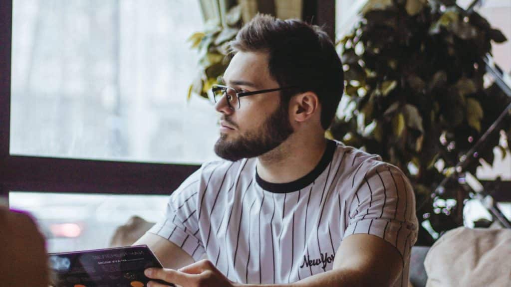 A man with a beard and glasses sits indoors looking out of a window.