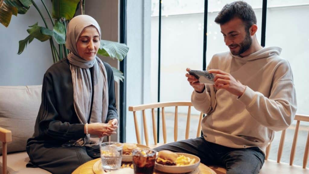 A man takes a photo of food with his phone while a woman watches.