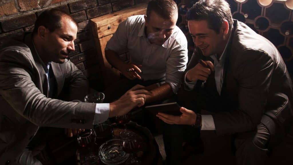 A group of men in suits smoking cigars and looking at a tablet in a dimly lit lounge.