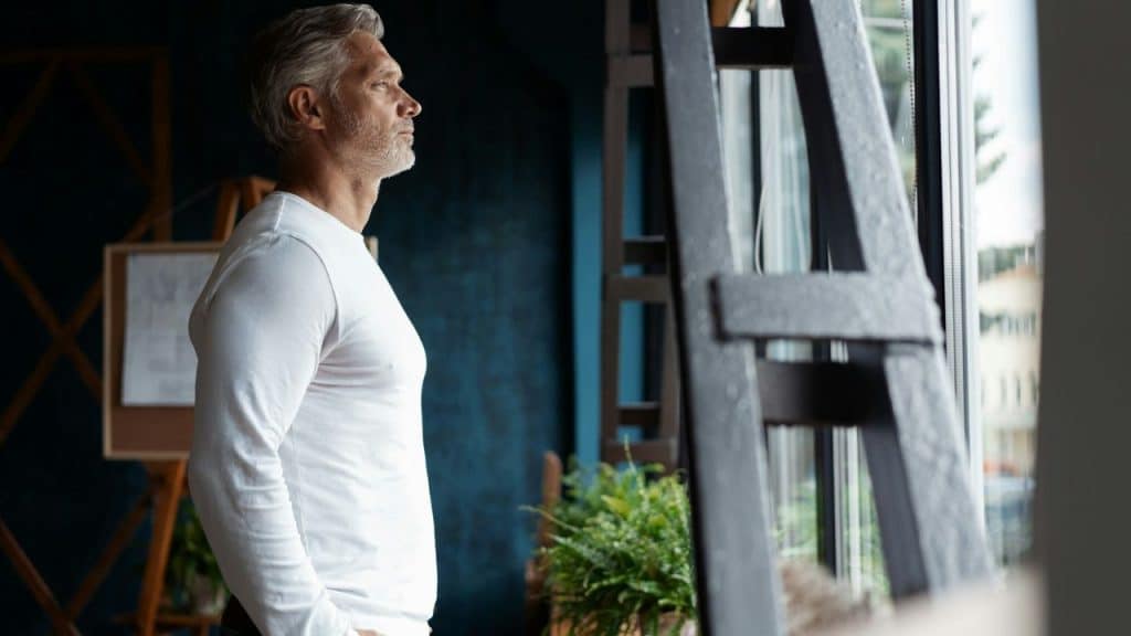 A middle-aged man in a white long-sleeved shirt stands indoors and looks out a window.