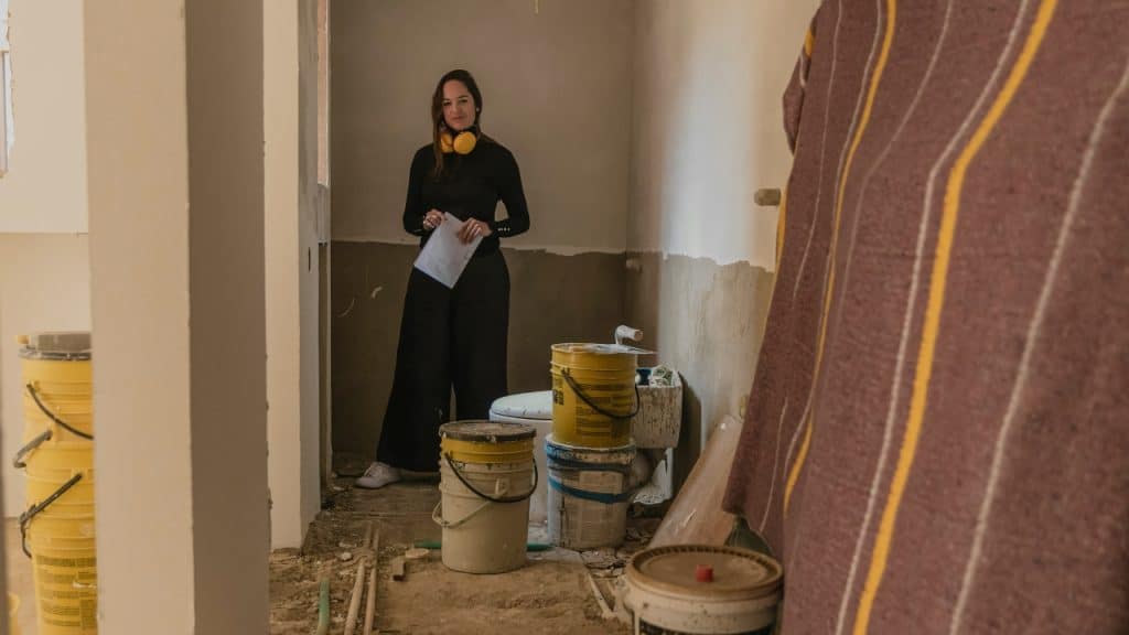 A woman stands in a room under construction surrounded by paint buckets and tools.