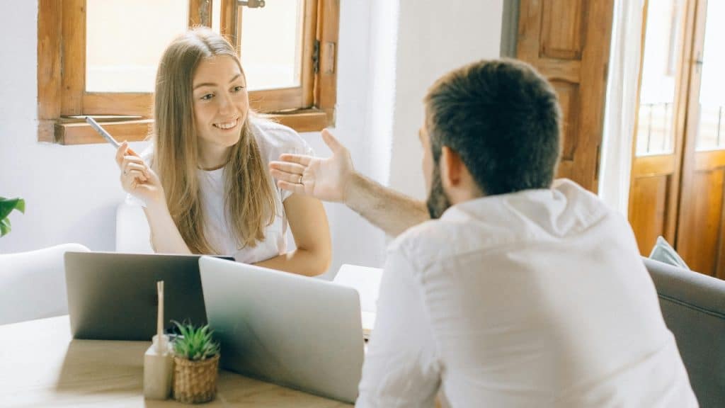 A man and a woman sit at a desk with laptops during a conversation.