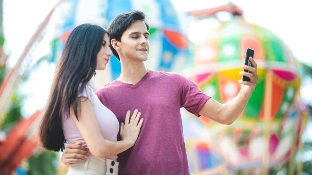 A man and woman pose for a selfie in front of a colorful carnival ride.