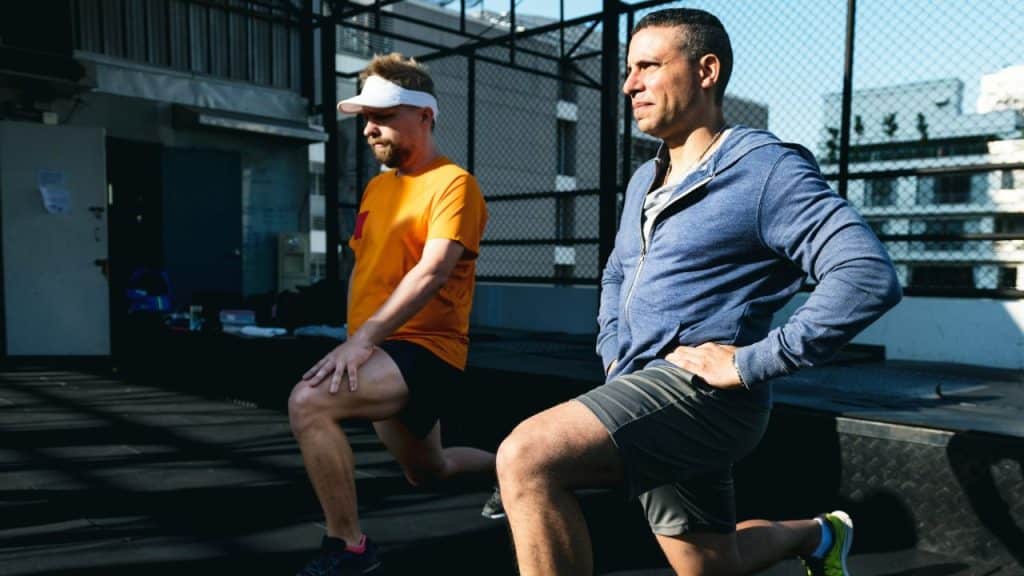 Two men in athletic wear performing lunges on an outdoor court during the day.