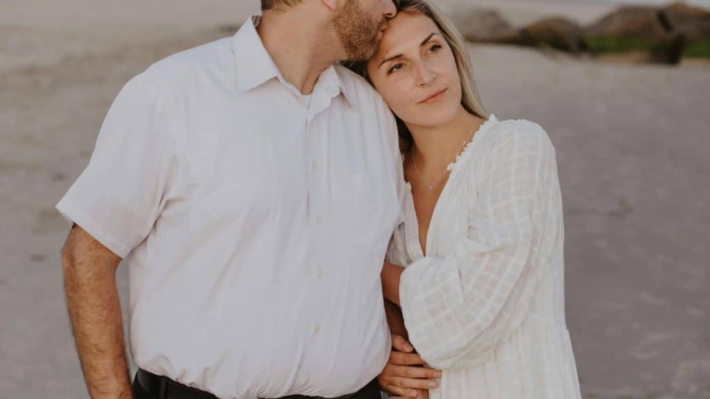 A man kissing a woman’s forehead as they stand together on a beach.