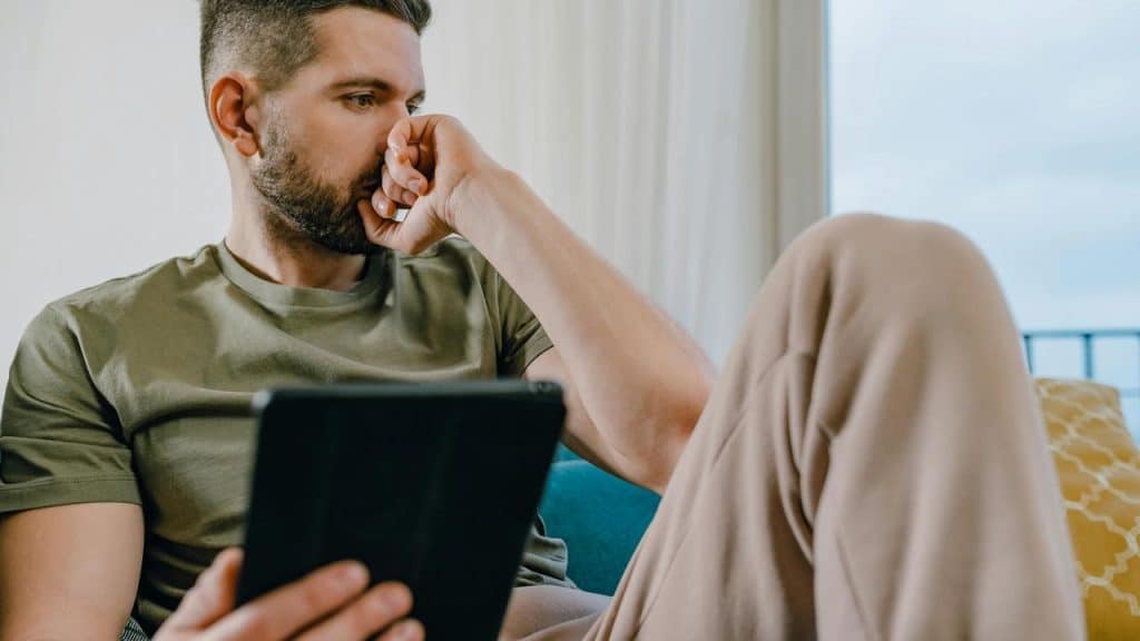 A man sitting on a couch, looking concerned while holding a tablet.