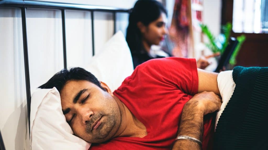 A person in a red short-sleeved top lying on a bed with a figure in dark clothing in the background.