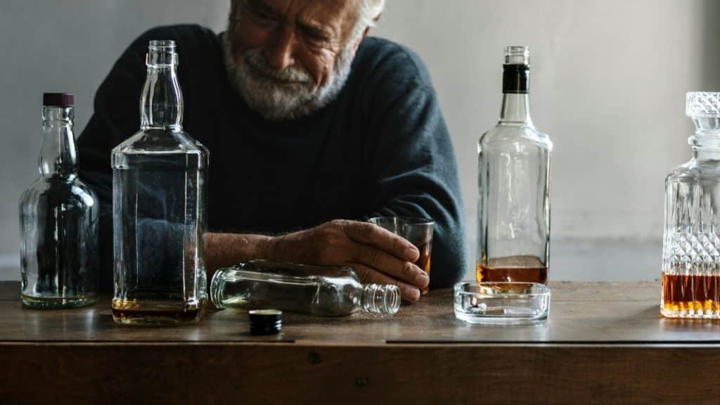 A man sits at a wooden table with several glass bottles, a glass, and an ashtray.