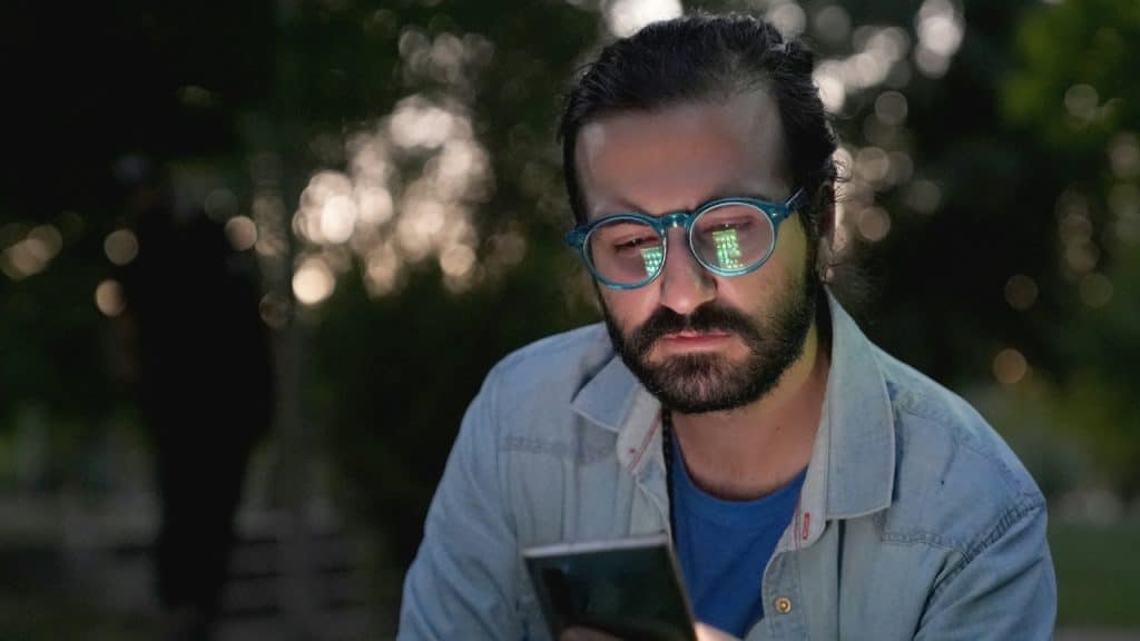 A man with a beard and glasses looks down at his glowing smartphone screen.