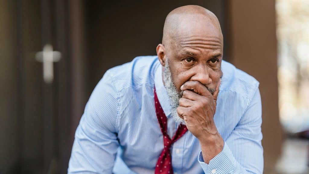 A man with a gray beard sits with his hand over his mouth looking concerned.