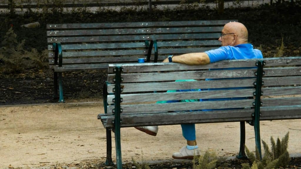 A man with glasses sits alone on a wooden park bench in a garden.