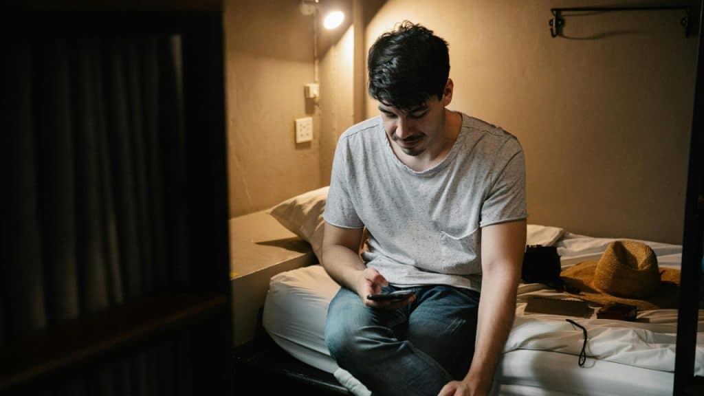 A man sits on a bed alone, looking down at his mobile phone screen.