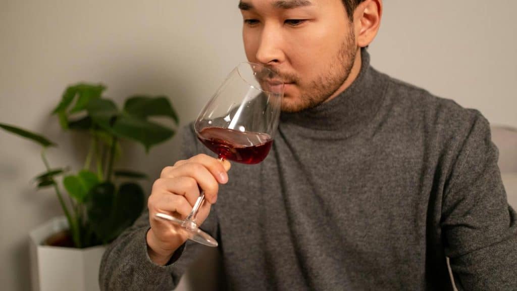 A man smelling a glass of red wine while sitting indoors.