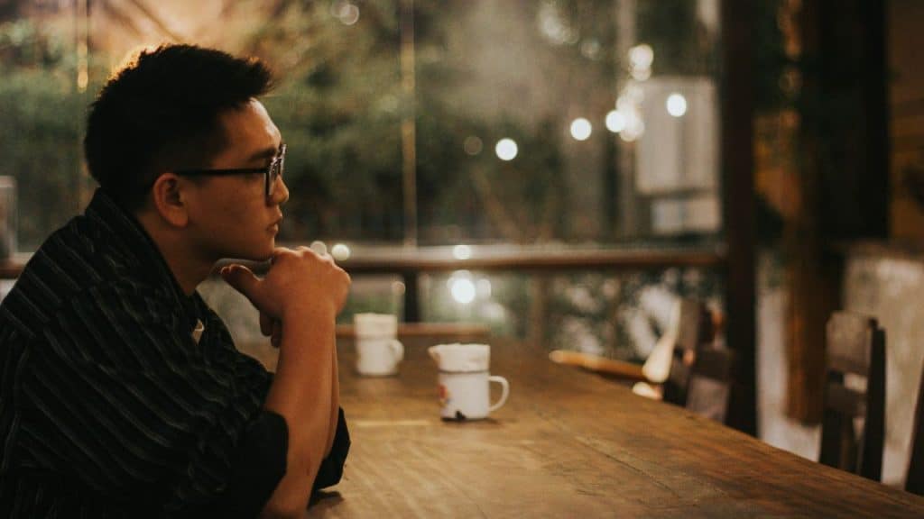 A man wearing glasses sits at a wooden table, resting his chin on his hand.