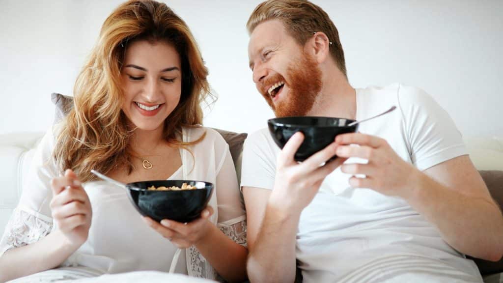 A man and a woman laugh together while holding bowls with spoons in them.