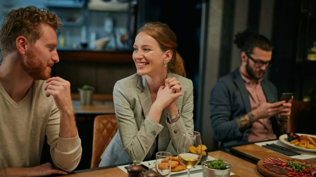 A smiling woman in a grey blazer sits at a dinner table talking to a man.