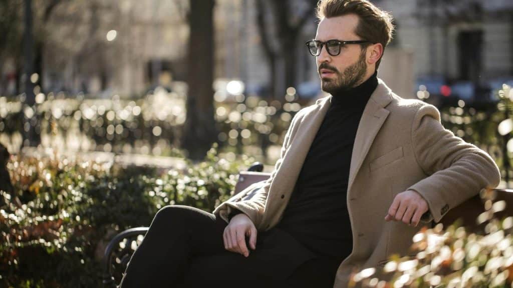 A stylish bearded man wearing glasses and a tan coat sitting on a park bench.