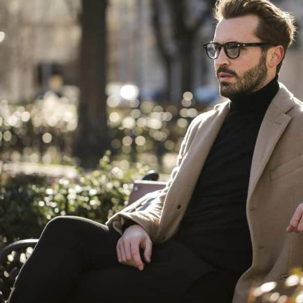 A stylish bearded man wearing glasses and a tan coat sitting on a park bench.