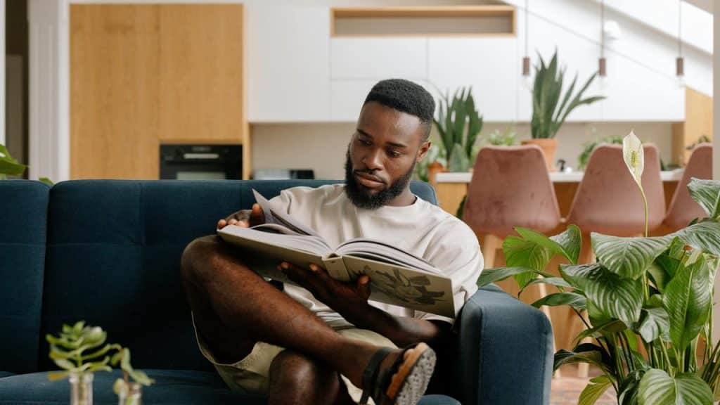 A man sitting on a couch reading a book in a bright living room.