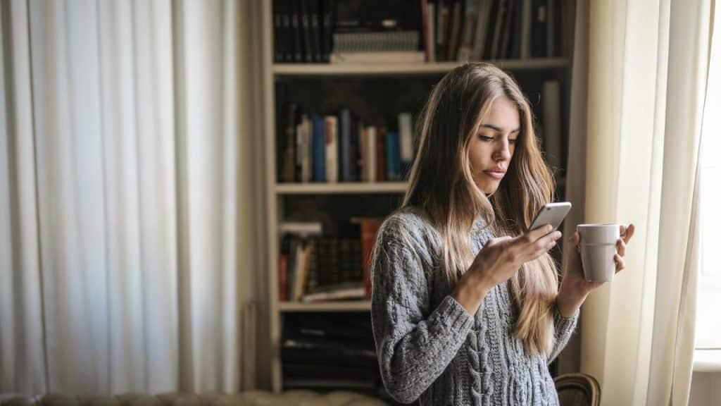 A woman holding a cup while looking at her smartphone indoors.