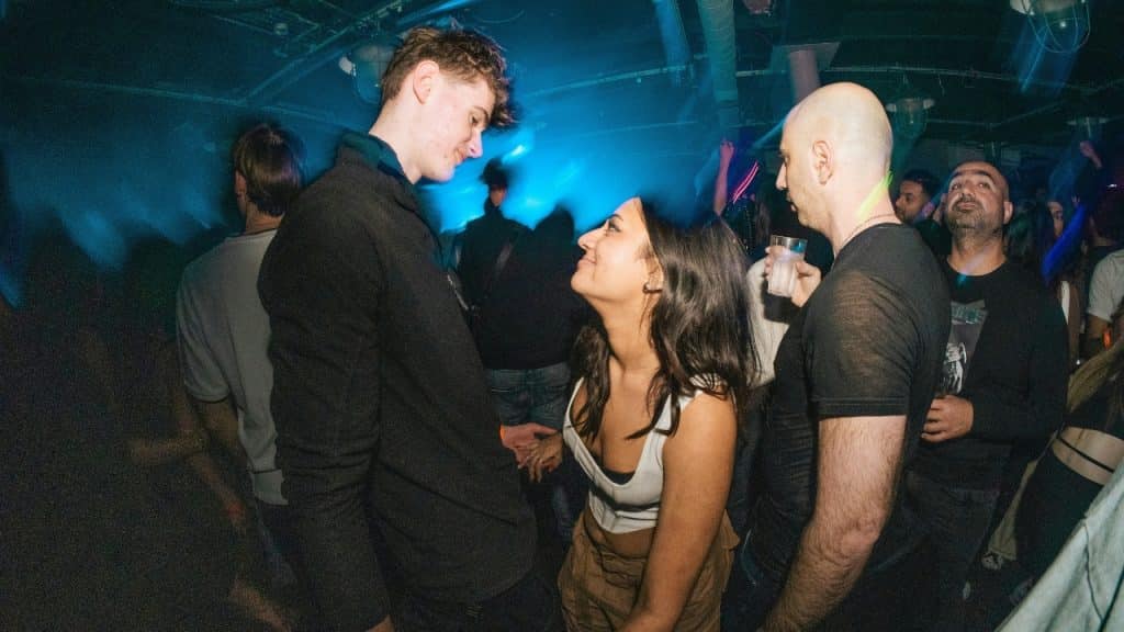 A man and woman stand close together in a dimly lit, crowded night club.