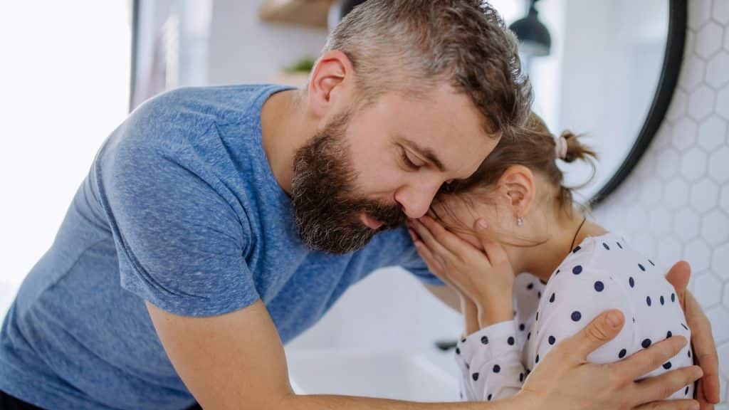 A man gently embraces a young girl who is covering her face with hands.