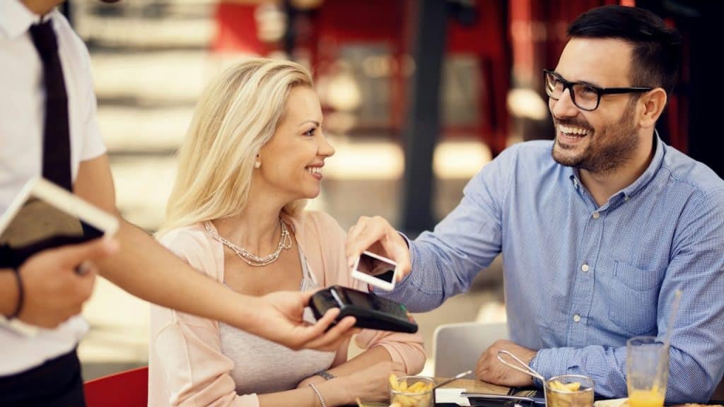 A man smiling at the waiter as he taps his phone on a cash machine, and a woman smiles at him.